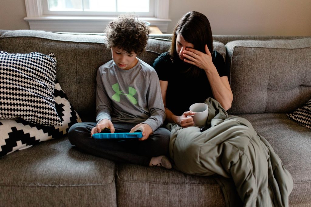 mother holding a coffee cup and watching son play on tablet while sitting on a couch