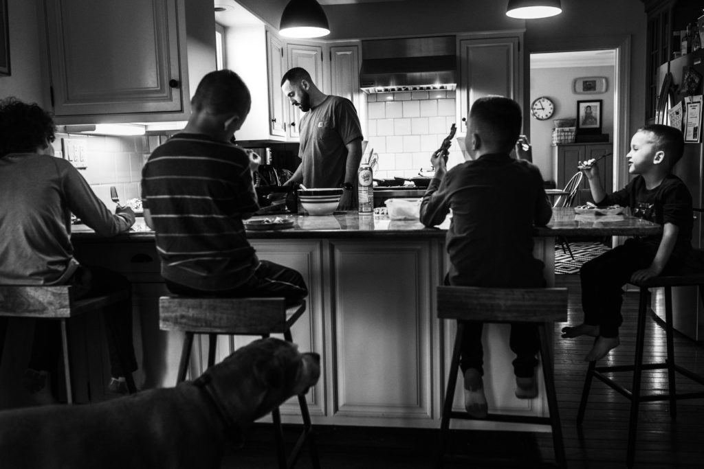 four children eating at a table while their father is in the kitchen