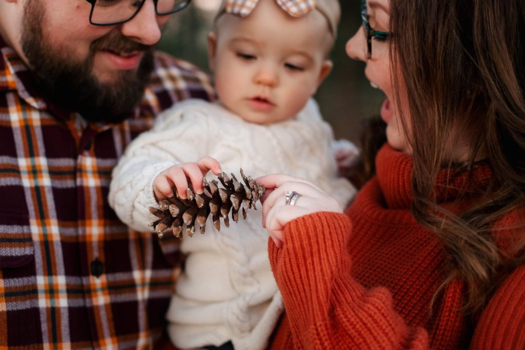parents smiling and holding their infant child between them while she is holding a pinecone