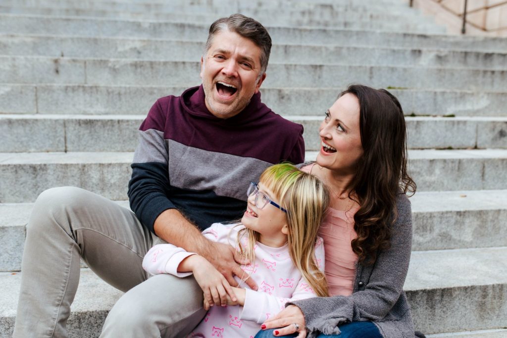 two parents sitting on steps laughing with their daughter in between them